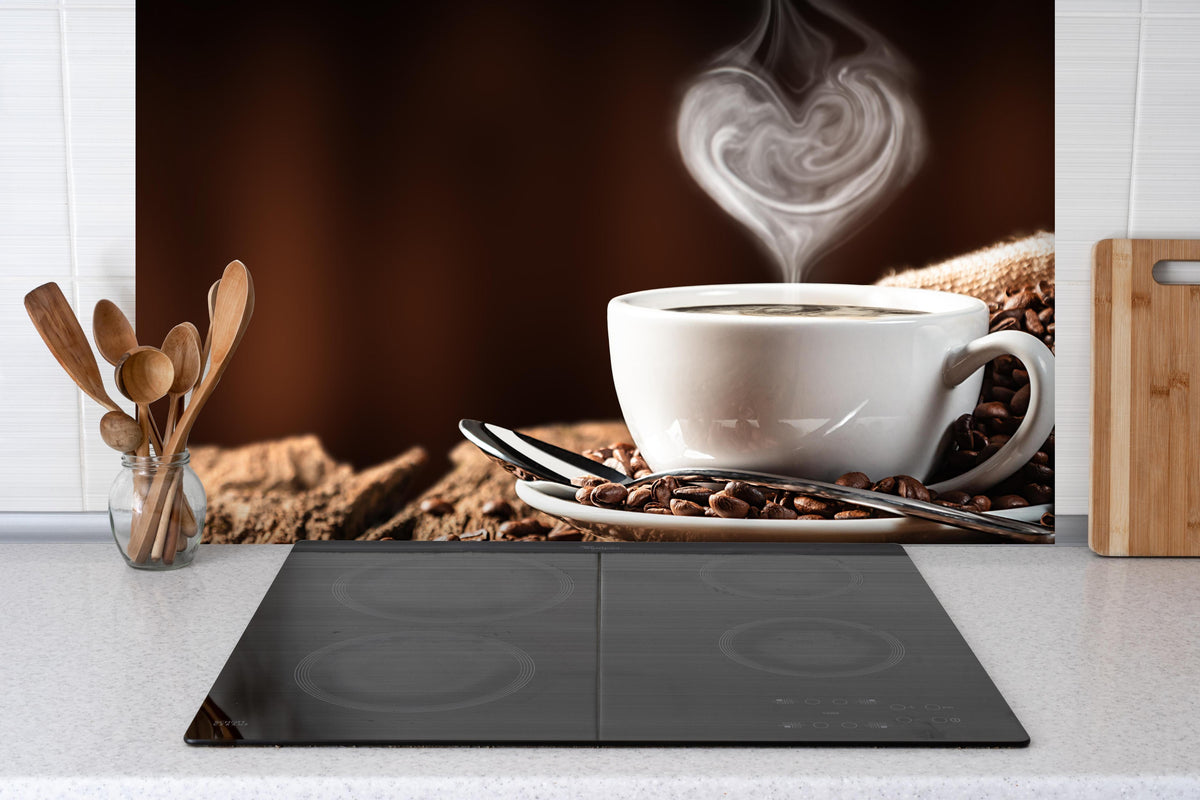 Spritzschutz - Kaffeetasse mit herzförmigem Dampf hinter einem Cerankochfeld zwischen Holz-Kochutensilien
