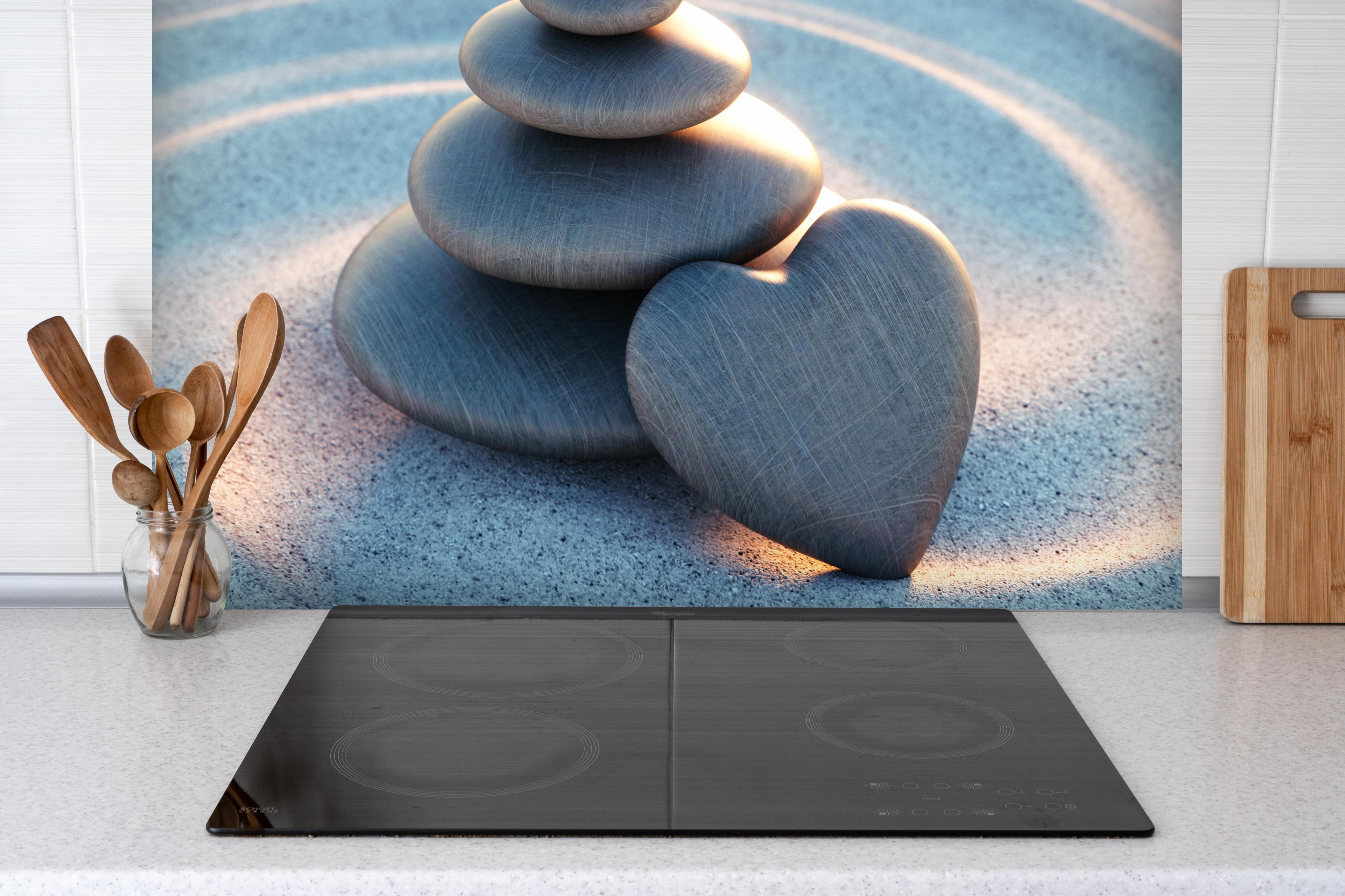 Spritzschutz - Liebevoller Turm aus Kieselsteinen hinter einem Cerankochfeld zwischen Holz-Kochutensilien
