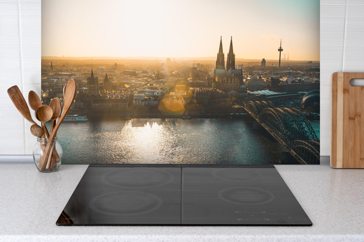 Spritzschutz - Panoramablick auf Köln bei Sonnenaufgang hinter einem Cerankochfeld zwischen Holz-Kochutensilien
