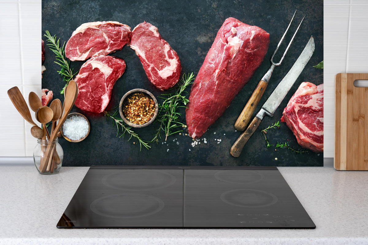 Spritzschutz - Steakvariationen auf Schieferplatte hinter einem Cerankochfeld zwischen Holz-Kochutensilien
