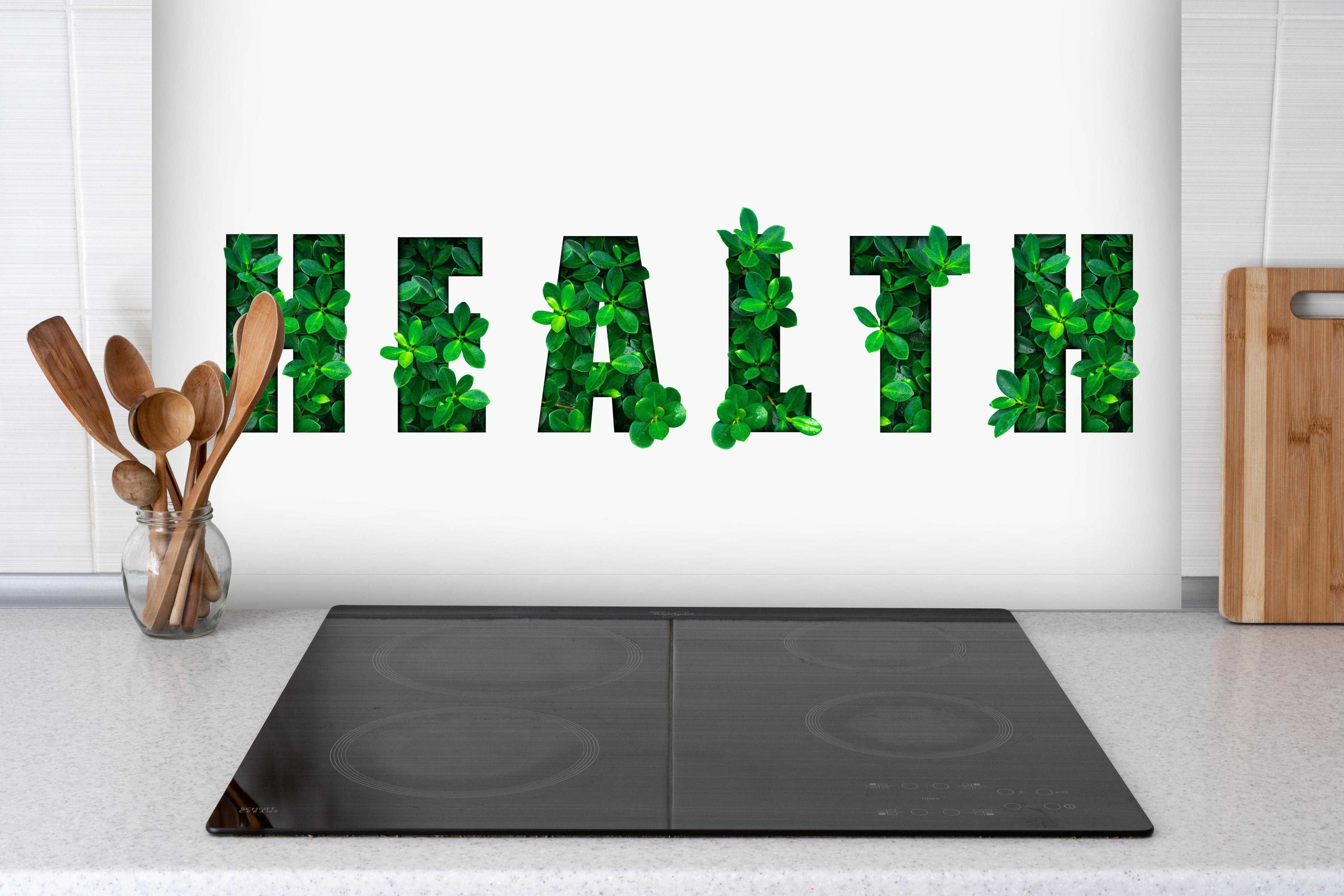 Spritzschutz - Wort HEALTH aus grünen Blättern hinter einem Cerankochfeld zwischen Holz-Kochutensilien
