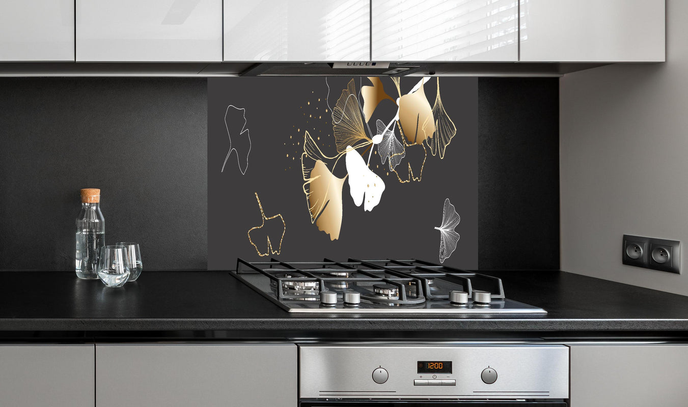 Spritzschutz - Stilisiertes Ginkgo-Design in Gold und Schwarz hinter einem Cerankochfeld zwischen Holz-Kochutensilien
