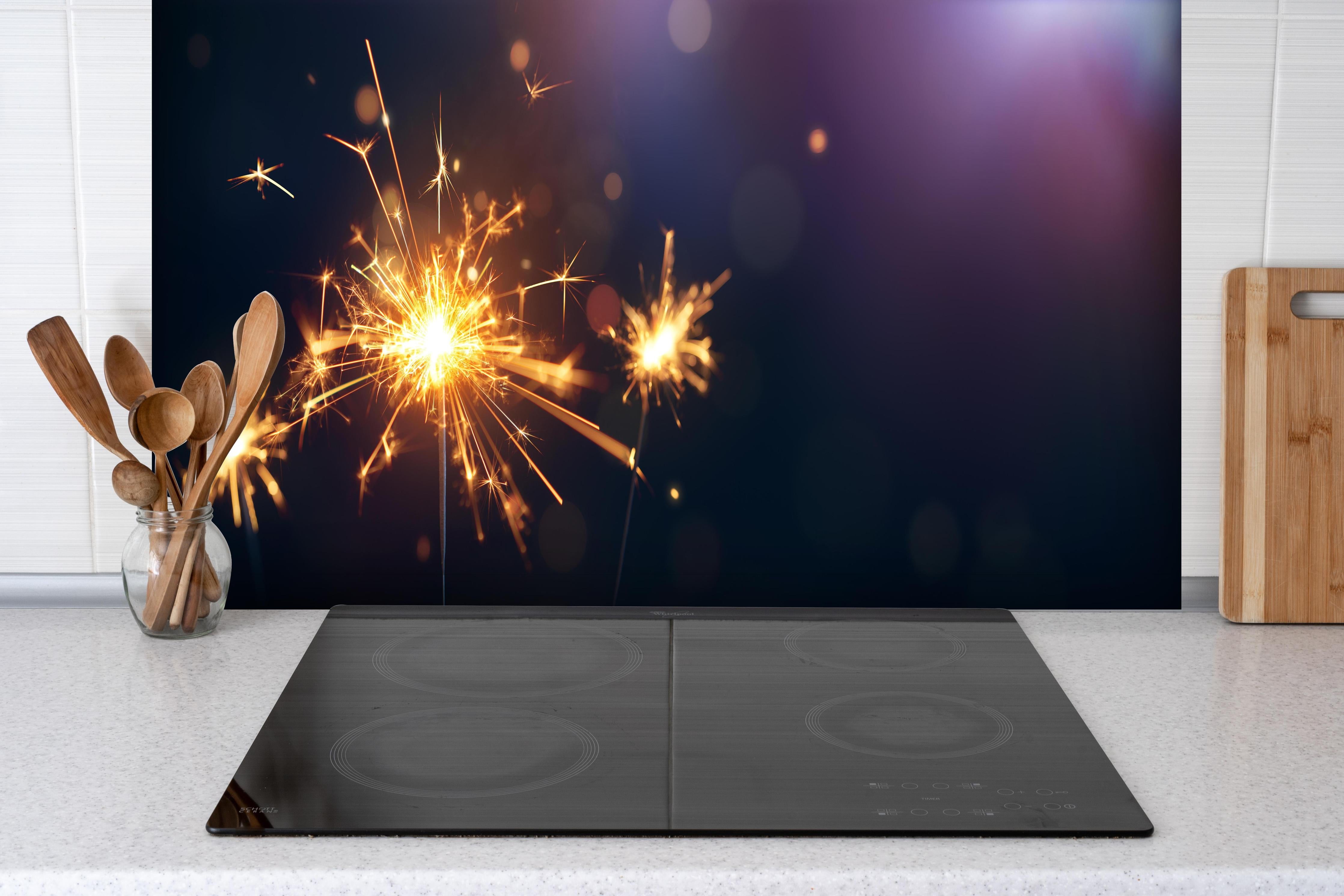 Spritzschutz - Strahlendes Wunderkerzen-Goldlichtfeuerwerk hinter einem Cerankochfeld zwischen Holz-Kochutensilien
