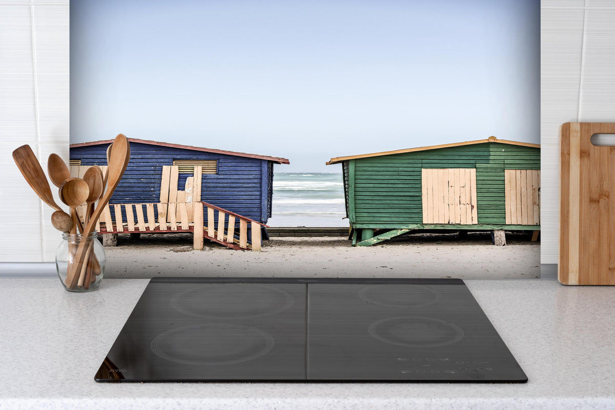 Spritzschutz - Strandhütten am Meer hinter einem Cerankochfeld zwischen Holz-Kochutensilien
