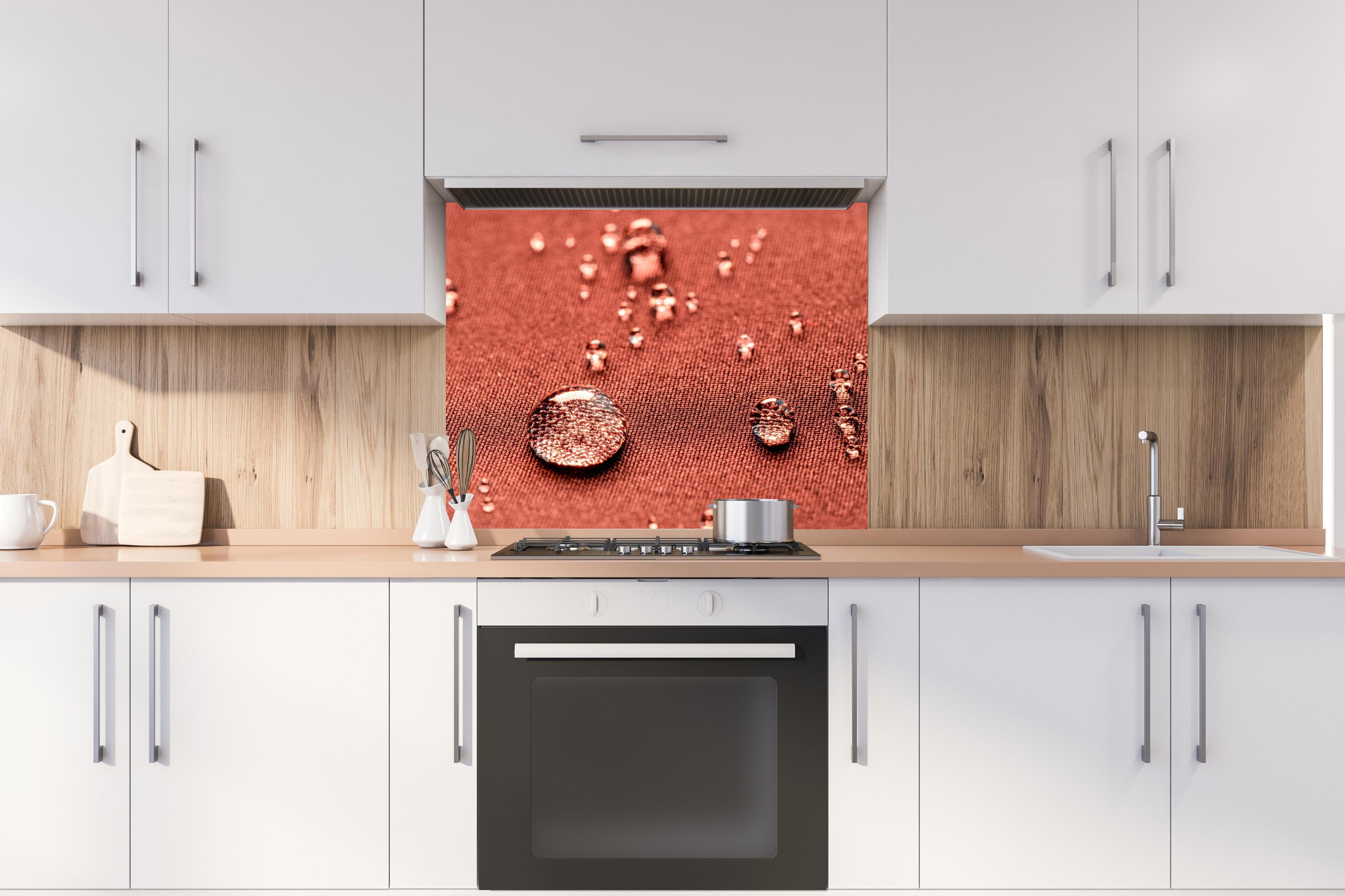 Spritzschutz - Wassertropfen auf rotem Stoff hinter einem Cerankochfeld zwischen Holz-Kochutensilien
