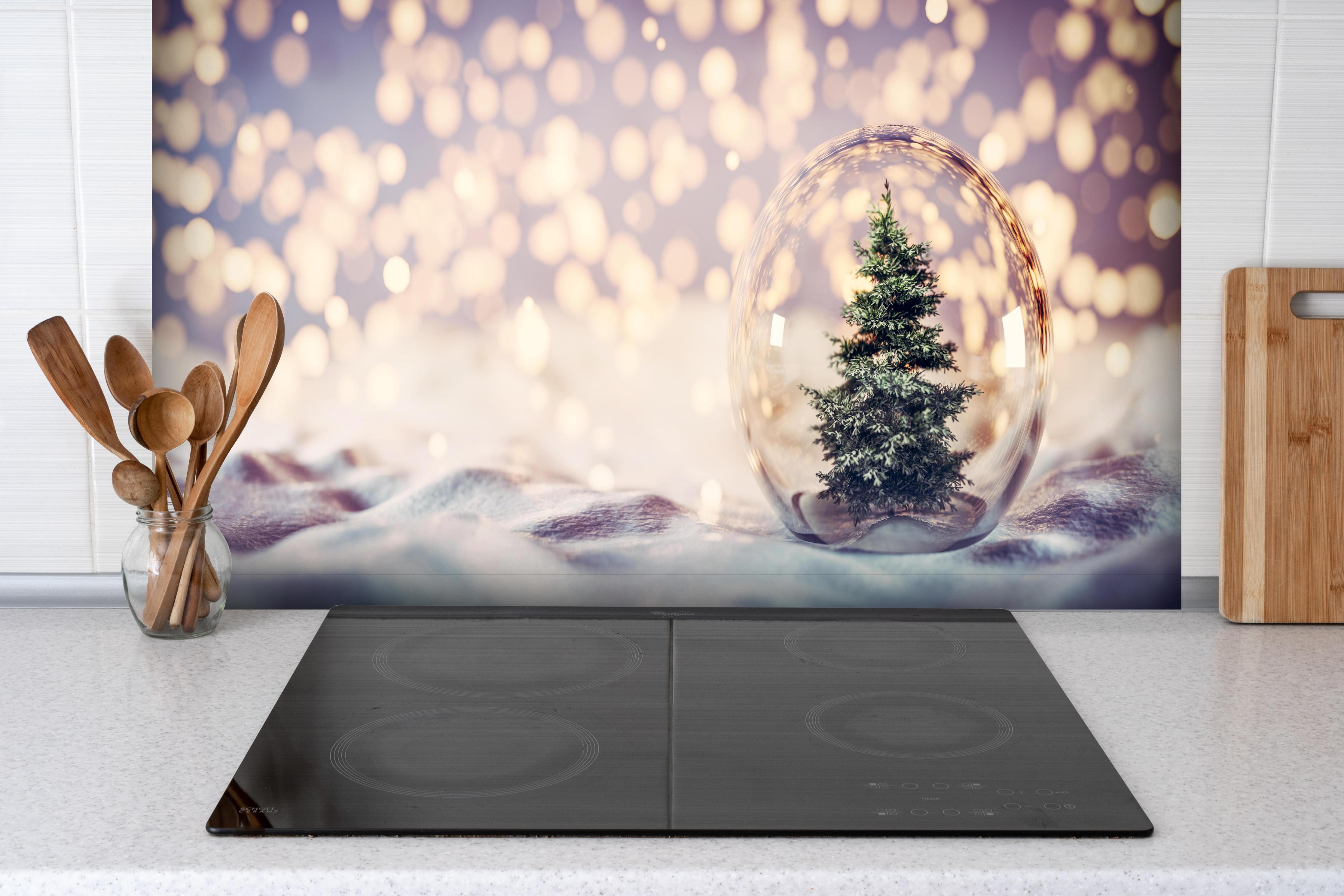 Spritzschutz - Weihnachtsbaum in einer Glaskugel hinter einem Cerankochfeld zwischen Holz-Kochutensilien
