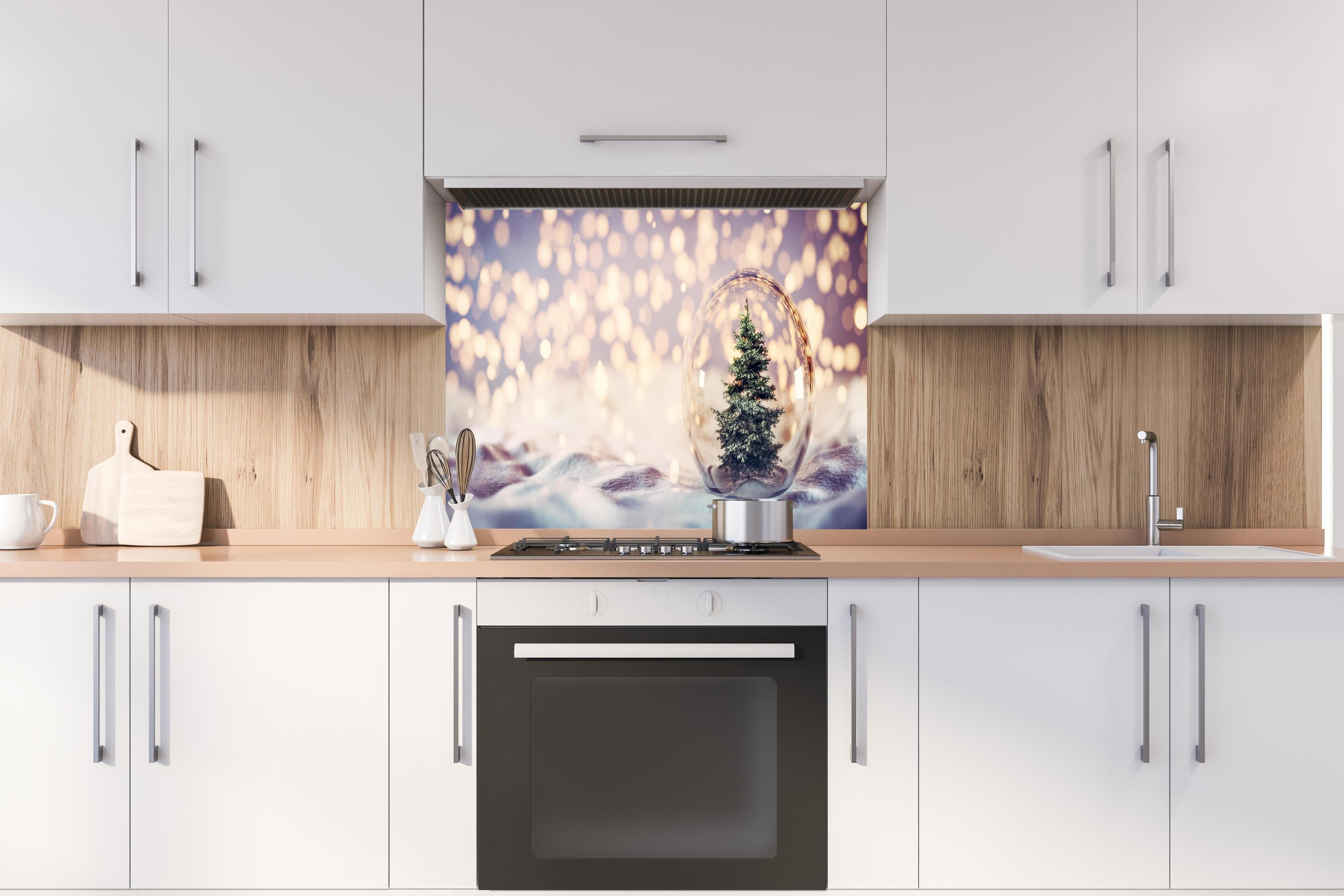 Spritzschutz - Weihnachtsbaum in einer Glaskugel hinter einem Cerankochfeld zwischen Holz-Kochutensilien
