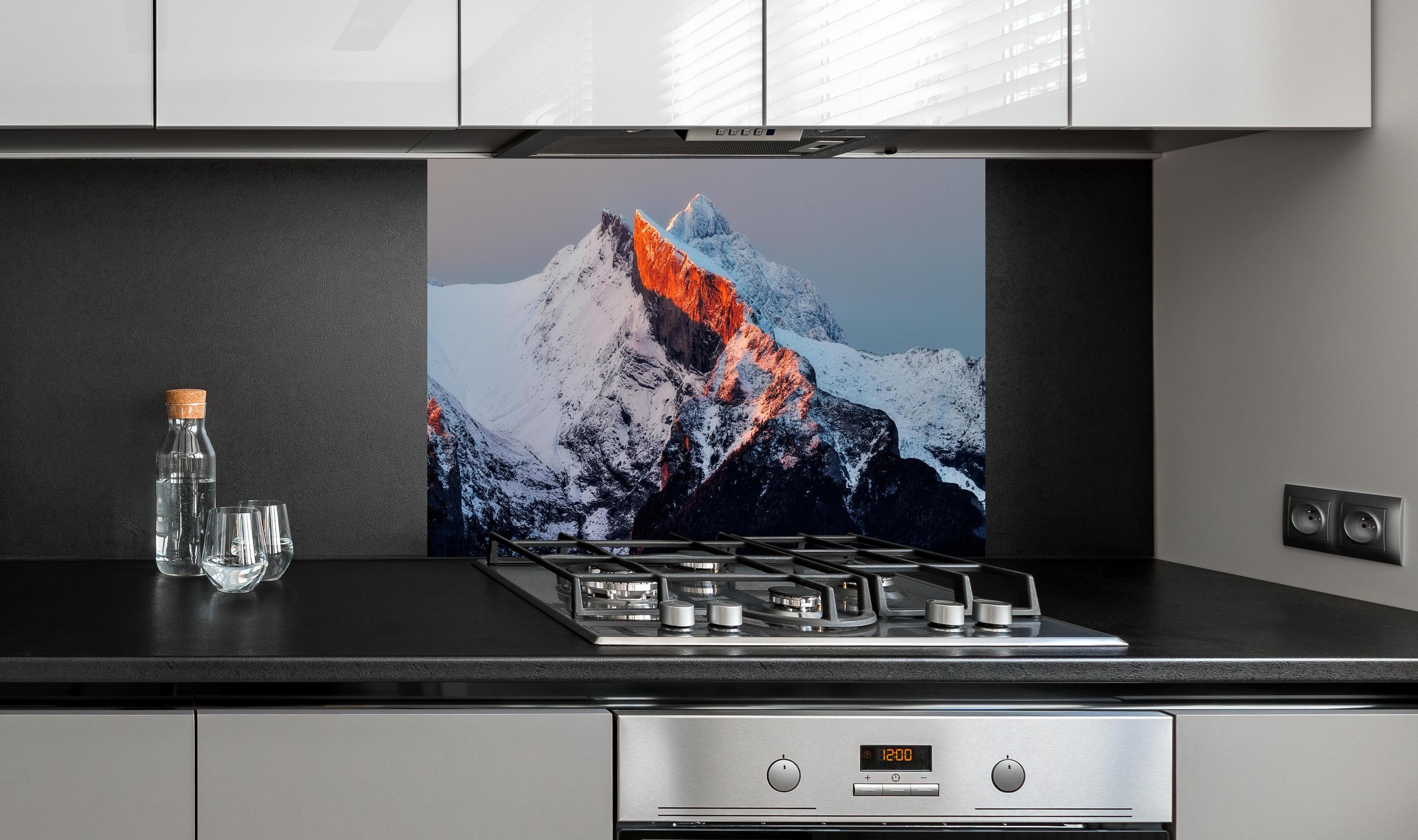 Spritzschutz - Winterlicher Sonnenaufgang über schneebedeckten Gipfeln hinter einem Cerankochfeld zwischen Holz-Kochutensilien
